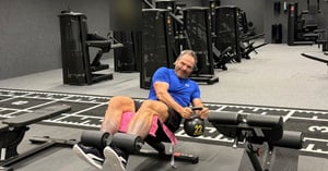 Igor Pauletič in a blue T-shirt on a gym bench, holding a kettlebell marked 22 and smiling at the camera.