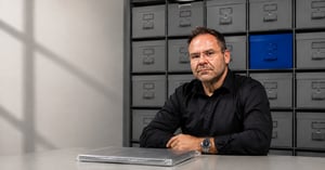Muškarac u crnoj košulji sjedi ispred zida sivih kutija za arhiviranje s jednom istaknutom plavom kutijom, a na stolu je zamotan predmet.