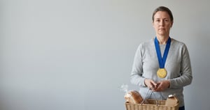 Ženska z medaljo okoli vratu drži pleteno košaro s kruhom in kartončkom proti svetlo sivi steni.